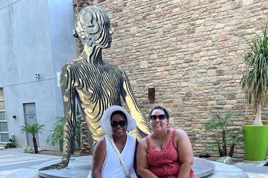 Two smiling women in sunglasses, one in a white sunhat, seated in front of a large layered metal human sculpture in an outdoor urban courtyard with a stone wall and potted palms.