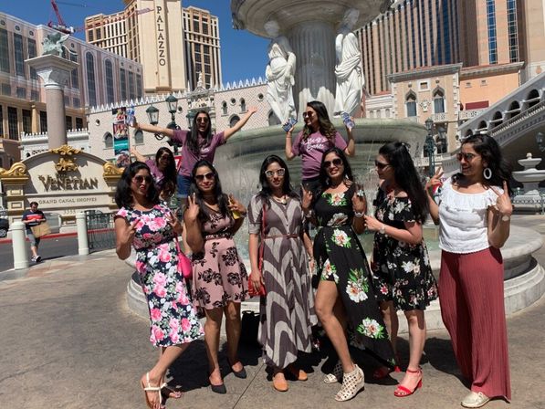 Cheerful group of nine women in summer dresses and sunglasses posing by a classical fountain outside an Italian‑style resort hotel on a sunny tourist promenade.