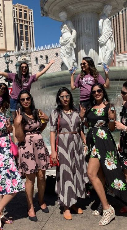 Cheerful group of nine women in summer dresses and sunglasses posing by a classical fountain outside an Italian‑style resort hotel on a sunny tourist promenade.