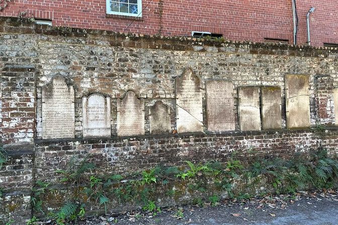 Row of weathered stone headstones set into a mossy flint-and-brick wall beneath a red-brick building, with ferns and a leaf-strewn path in front.