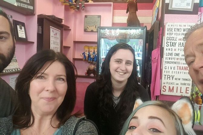 Group selfie of five smiling people inside a pink-themed boutique or gift shop, one wearing a cat-ear headband, with decorative wall signs, shelves of toys and a refrigerated display in the background.