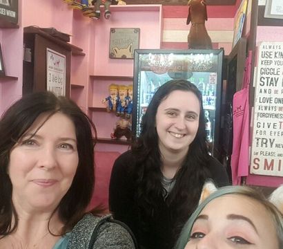 Group selfie of five smiling people inside a pink-themed boutique or gift shop, one wearing a cat-ear headband, with decorative wall signs, shelves of toys and a refrigerated display in the background.