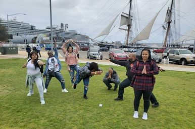 Eight people striking playful yoga and martial-arts poses on a waterfront lawn with a docked cruise ship and a tall sailing ship in the harbor under a cloudy sky.