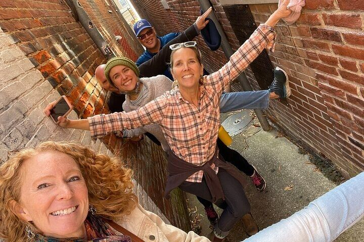 Playful group selfie of five friends posing in a narrow downtown brick alleyway, arms outstretched against the walls and smiling.