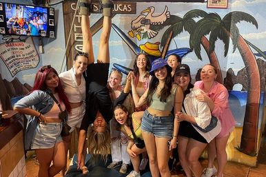 Group of friends laughing and posing inside a lively Cabo San Lucas beach bar with a colorful palm-tree and pelican mural; one person hanging upside down while others cheer.