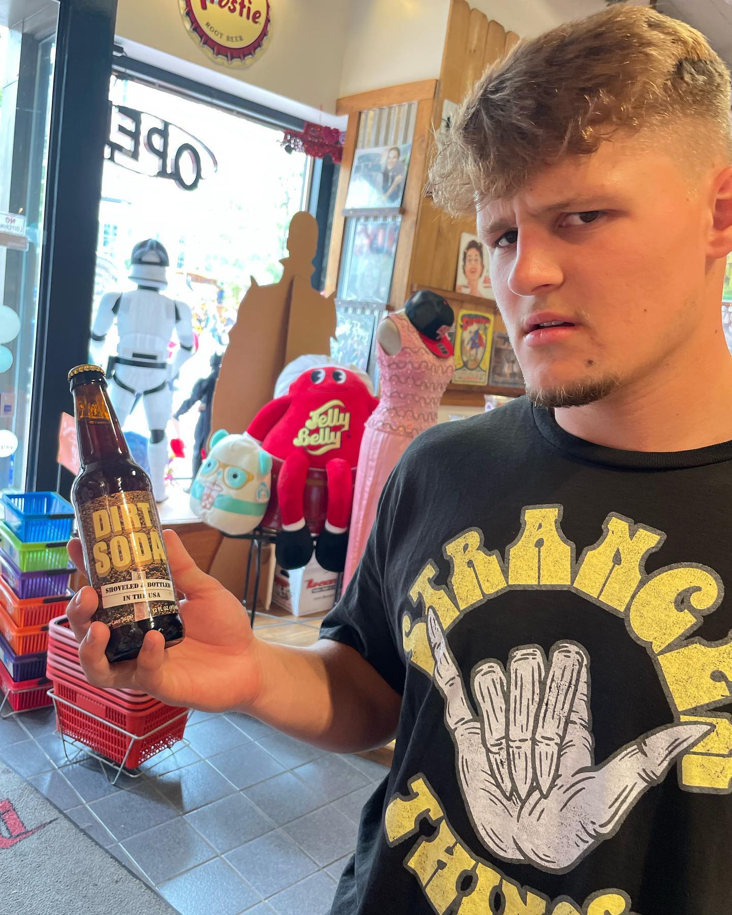 Young man in a graphic tee holding a bottle labeled "Dirt Soda" inside a quirky novelty gift shop with Jelly Belly plush, costume mannequin, and a Stormtrooper figure by the open door.