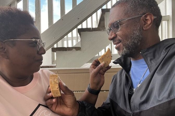 Two adults playfully sharing caramel candy on a sunlit outdoor wooden porch with a staircase and railings in the background.