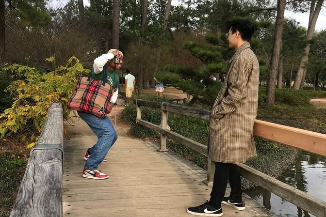 Two people on a wooden footbridge in a park by a pond — one squatting to take a photo holding a plaid tote and wearing red sneakers, the other leaning in a long checkered coat amid pine trees and autumn foliage.
