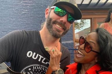Playful moment on an outdoor urban patio — bearded man in a cap and mirrored sunglasses smiling while a laughing woman in a red jacket and dark sunglasses playfully tucks a dollar bill into his shirt.
