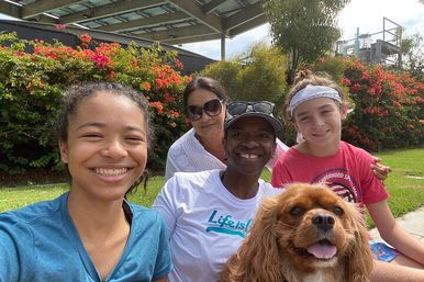 Smiling group selfie of four people and a happy brown dog sitting on grass in a sunny park with red flowering bushes and a solar-panel pergola overhead.
