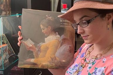 Person wearing a hat and glasses holding a shrink-wrapped 2020 wall calendar featuring a classical painting of a seated girl reading, displayed on a shop shelf.