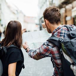 Two backpackers consulting a paper map and pointing on a cobblestone city street — urban sightseeing and travel