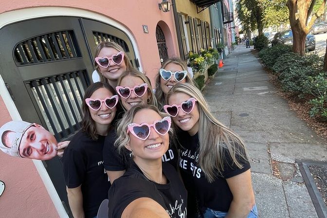 Six women in matching black shirts wearing pink and white heart-shaped sunglasses, smiling for a selfie on a sunny sidewalk in front of a pastel pink row house, one holding a cardboard face cutout.