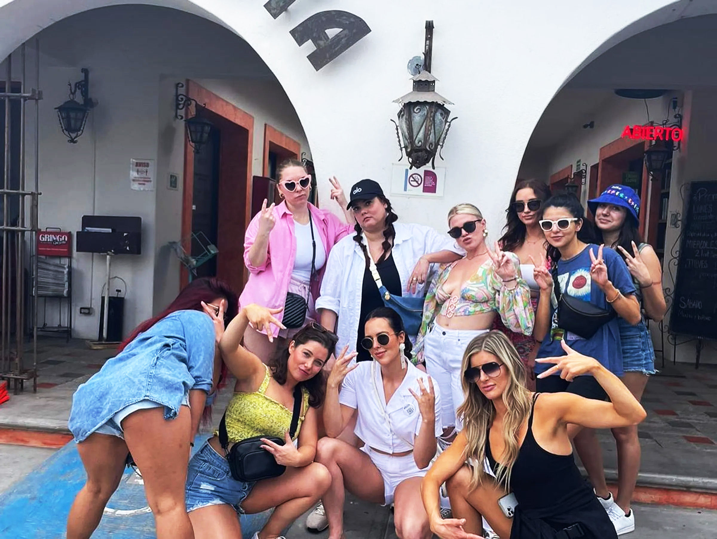Playful group of women in colorful summer outfits and sunglasses posing with peace signs under white stucco arches at a sunny courtyard, giving a fun vacation vibe.
