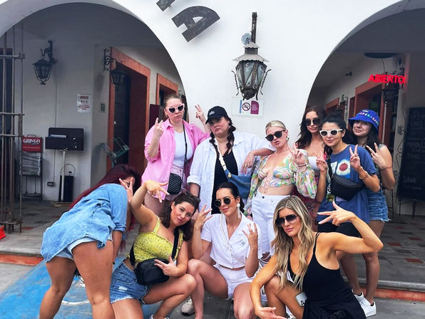 Playful group of women in colorful summer outfits and sunglasses posing with peace signs under white stucco arches at a sunny courtyard, giving a fun vacation vibe.