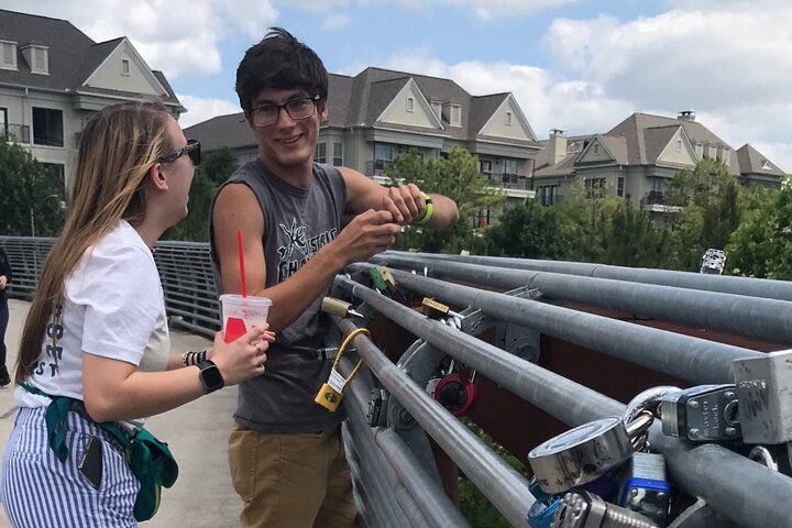 Playful pair attaching padlocks to a pedestrian bridge railing covered in love locks, one holding a drink, with urban apartment buildings and trees behind them under a cloudy sky.