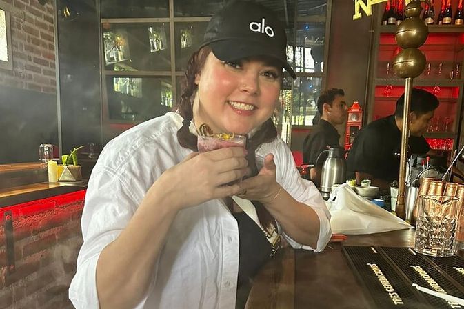 Smiling woman in a black cap and white shirt holding a colorful cocktail at an urban bar with exposed brick, red accent lighting, glassware, and bartenders in the background.