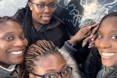 Close-up selfie of four smiling women by a large map display, one pointing to North America — fun travel photo at an outdoor map monument