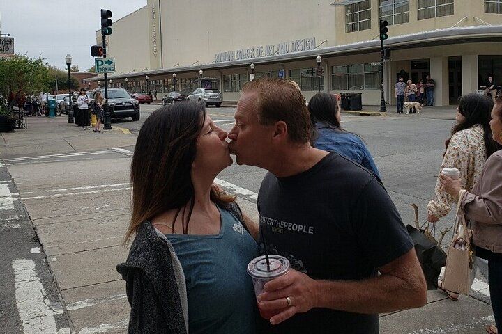 Couple sharing a kiss on a downtown crosswalk, man holding an iced drink, with pedestrians, parked cars and a large beige city building in the background.