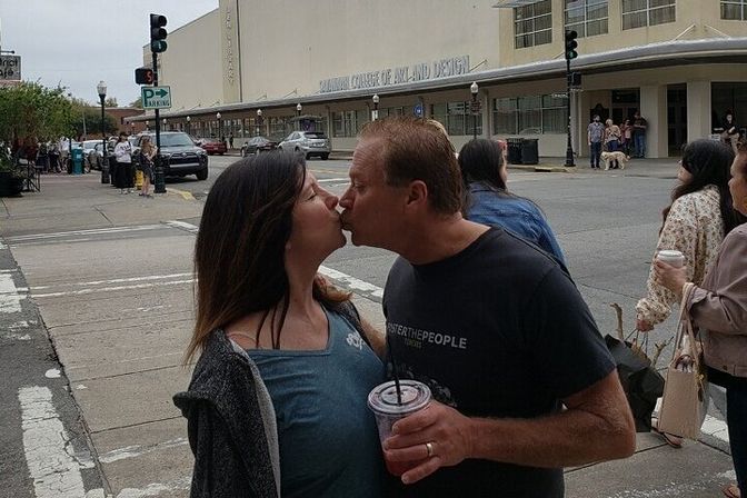 Couple sharing a kiss on a downtown crosswalk, man holding an iced drink, with pedestrians, parked cars and a large beige city building in the background.