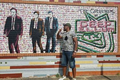 Man in camo shirt and denim shorts striking a playful pose next to a colorful Deep Ellum street mural on a brick wall featuring three suit-wearing silhouettes and neon-style 'Deep Ellum TX' graffiti.