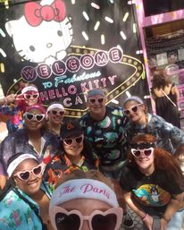 Group selfie of eight people wearing 'The Party' headbands and pink heart-shaped sunglasses, smiling in front of a pink-themed café storefront with a large white cartoon cat, neon 'Welcome' sign and colorful sprinkle pattern.