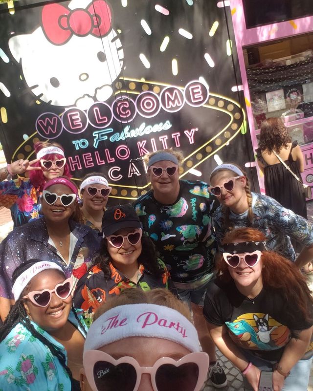 Group selfie of eight people wearing 'The Party' headbands and pink heart-shaped sunglasses, smiling in front of a pink-themed café storefront with a large white cartoon cat, neon 'Welcome' sign and colorful sprinkle pattern.