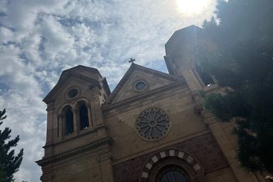 Sunlit historic sandstone church facade with twin bell towers, ornate circular rose window and cross against a partly cloudy sky, framed by evergreen trees.