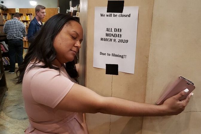Person hugging a pillar inside a bookstore, eyes closed and holding a smartphone, beside a taped sign reading "We will be closed ALL DAY MONDAY MARCH 9, 2020 Due to filming," with bookshelves and shoppers in the background.