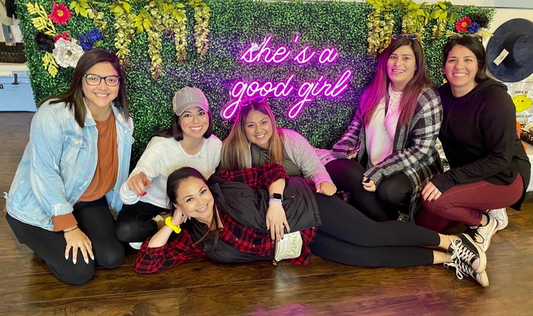 Six smiling women in casual outfits pose on a wood floor in front of a faux-greenery photo wall with a pink neon sign reading 'she's a good girl'; one woman lounges in front while the others kneel around her in a playful group shot.