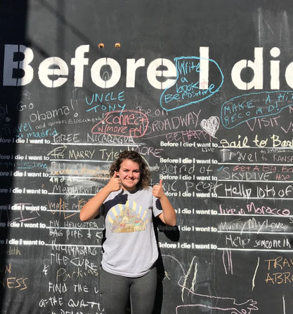 Smiling young woman giving two thumbs up in front of a sunlit public "Before I die" chalkboard wall covered in colorful handwritten bucket-list messages.