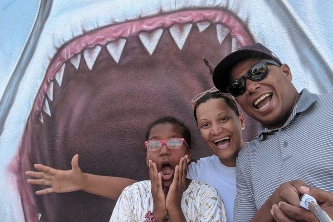 Family selfie in front of a giant shark street mural, child with pink glasses making a shocked pose and two adults laughing while holding a selfie stick.