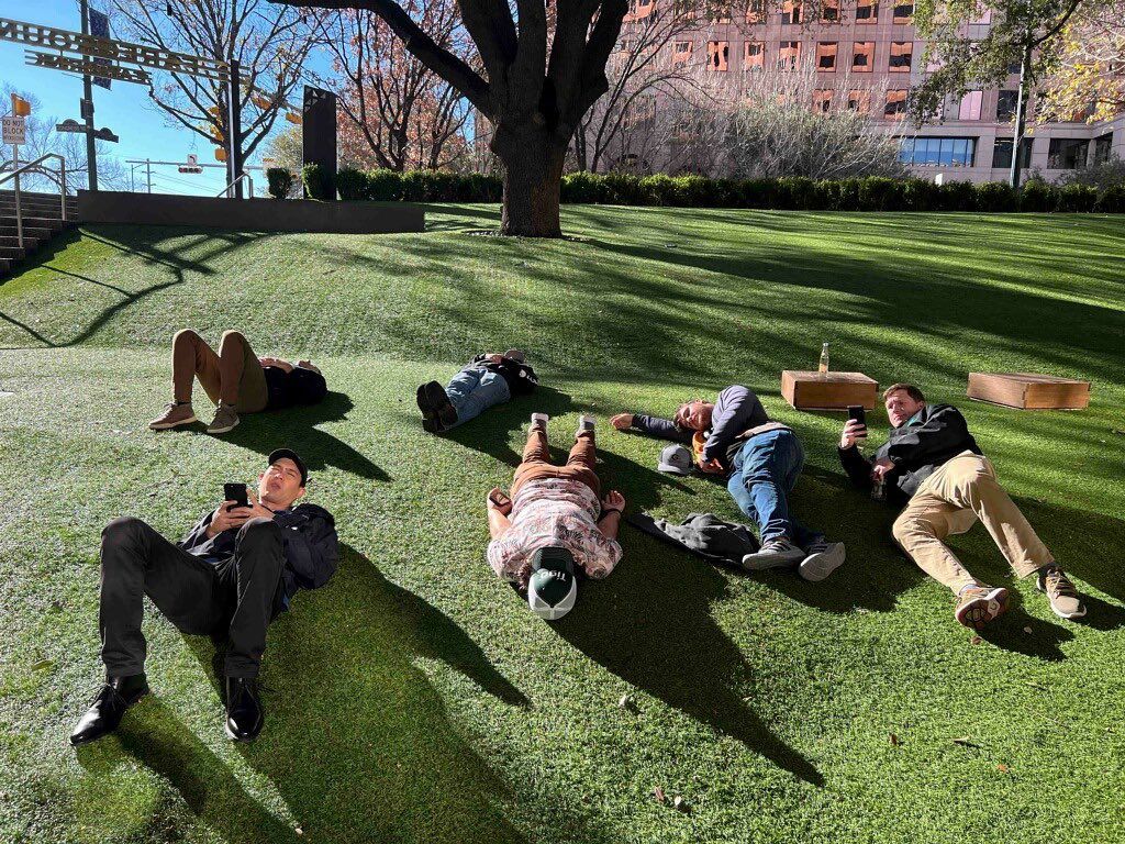 Six people lying on a sunlit urban park lawn on a grassy slope beneath a large tree, some checking phones, with long shadows and office buildings in the background.