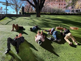Six people relaxing on a sunny downtown park lawn, lying on a sloped green turf beneath a large tree, some using phones with city buildings and steps in the background.