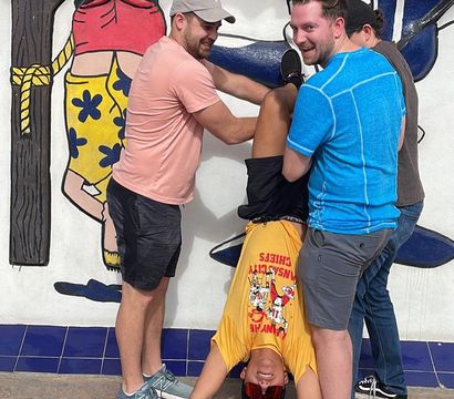 Three men laugh as they hold a fourth friend upside down in front of a colorful outdoor mural above a blue tiled base, playful group pose