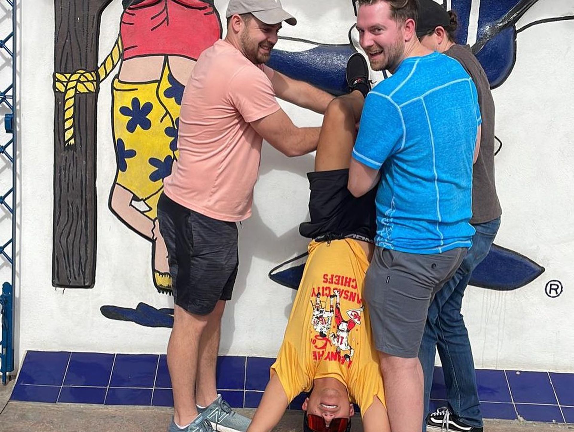 Three men laugh as they hold a fourth friend upside down in front of a colorful outdoor mural above a blue tiled base, playful group pose