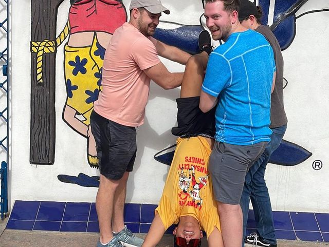 Three men laugh as they hold a fourth friend upside down in front of a colorful outdoor mural above a blue tiled base, playful group pose