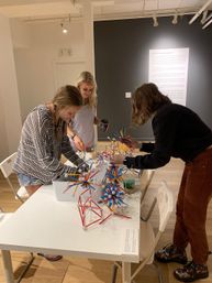 Three people at a gallery-style workshop building colorful geometric sculptures from red, blue and yellow rods and white connector balls on a white table, minimalist gallery wall in the background.
