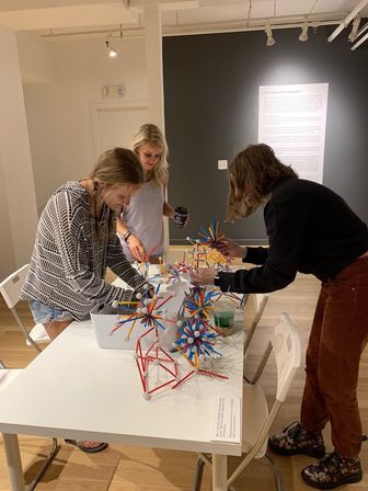 Three people at a gallery-style workshop building colorful geometric sculptures from red, blue and yellow rods and white connector balls on a white table, minimalist gallery wall in the background.