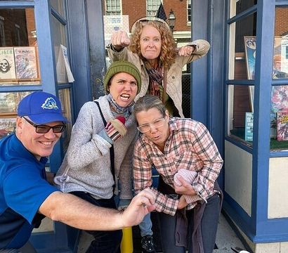 Group of four adults striking playful mock-angry poses at the entrance of a blue-fronted comic book shop, with comic books visible in the window display