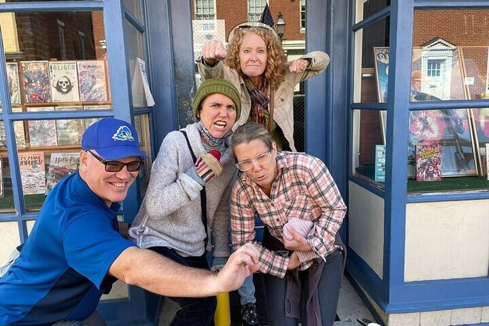 Group of four adults striking playful mock-angry poses at the entrance of a blue-fronted comic book shop, with comic books visible in the window display