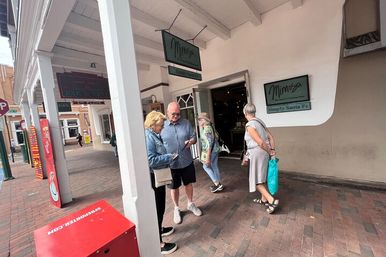 Three shoppers chatting outside a white-walled storefront with green signs under a covered, brick-paved historic Santa Fe shopping arcade
