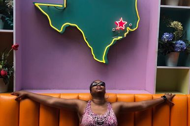 Person relaxing with arms outstretched on an orange booth beneath a neon-outlined Texas map with a glowing red star, purple wall backdrop and potted flowers.
