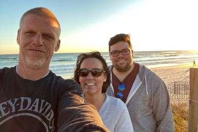 Three people smiling for a selfie on a coastal beach at sunset, waves rolling onto a sandy shore, one wearing sunglasses and another in a hoodie.