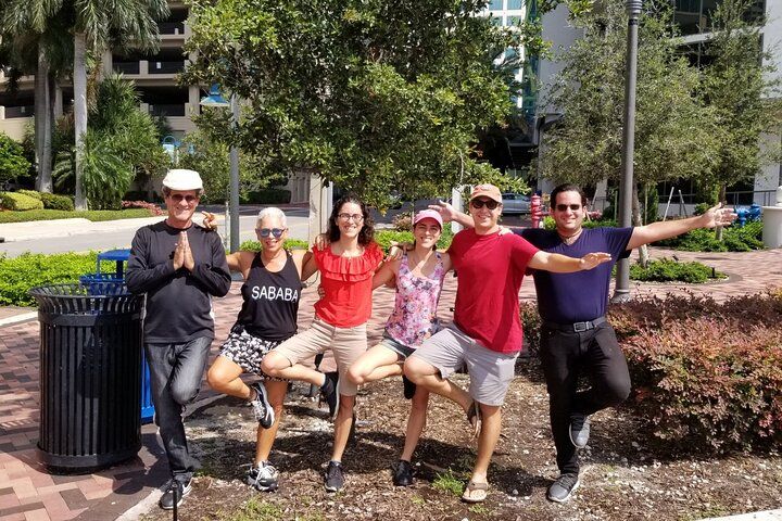 Six adults balancing in yoga tree pose, smiling and linking arms in a sunny downtown park with palm trees, paved walkway and buildings in the background.