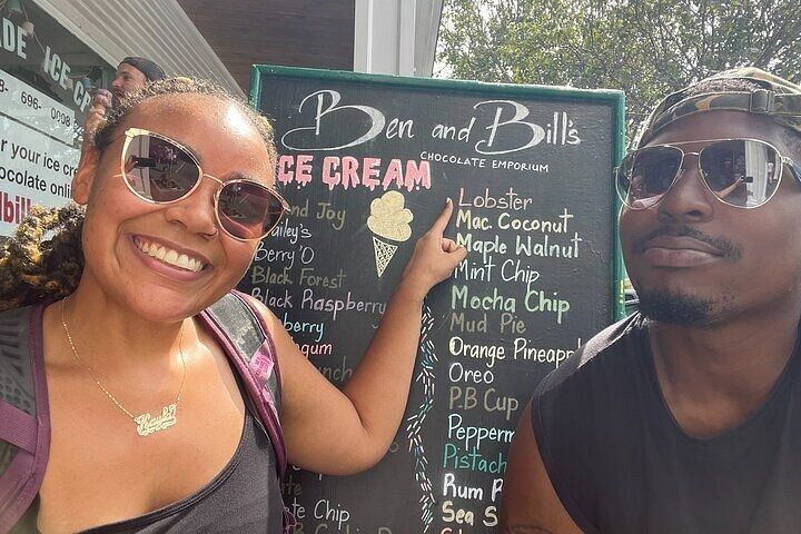 Two smiling people in sunglasses pose by an outdoor ice cream stand’s chalkboard menu listing playful flavors like lobster, macadamia coconut and mint chip.