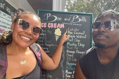 Two smiling people in sunglasses pose by an outdoor ice cream stand’s chalkboard menu listing playful flavors like lobster, macadamia coconut and mint chip.