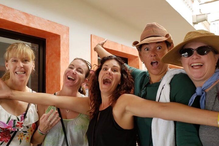 Five laughing women friends posing with arms outstretched in a sunlit courtyard, wearing casual summer clothes, hats and sunglasses — playful vacation snapshot.