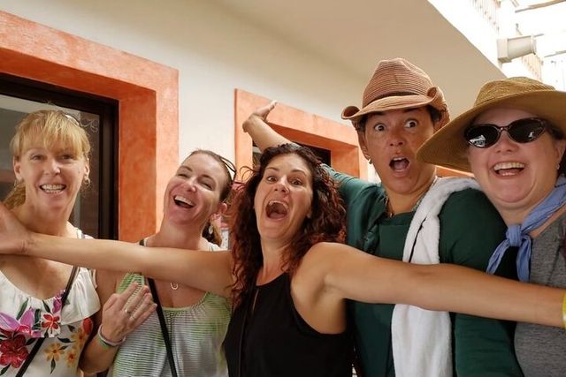 Five laughing women friends posing with arms outstretched in a sunlit courtyard, wearing casual summer clothes, hats and sunglasses — playful vacation snapshot.