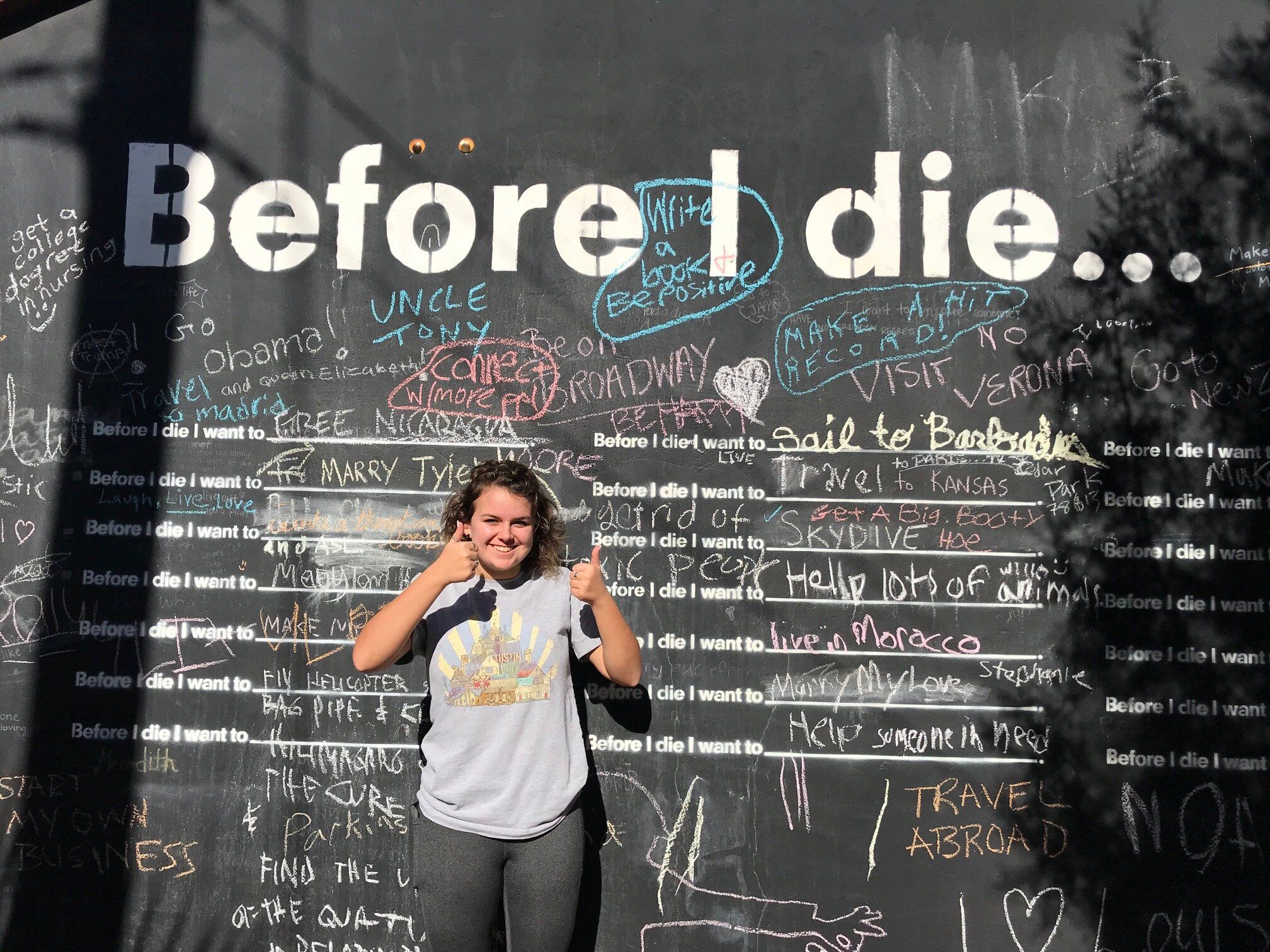 Smiling person giving thumbs-up in front of a large outdoor "Before I die..." interactive chalkboard wall filled with colorful handwritten wishes and messages.